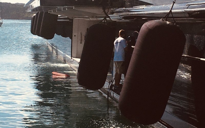 A crew member of a mega yacht stands on the exterior lower deck, performing maintenance work, while the yacht's massive black fenders are visible at the front