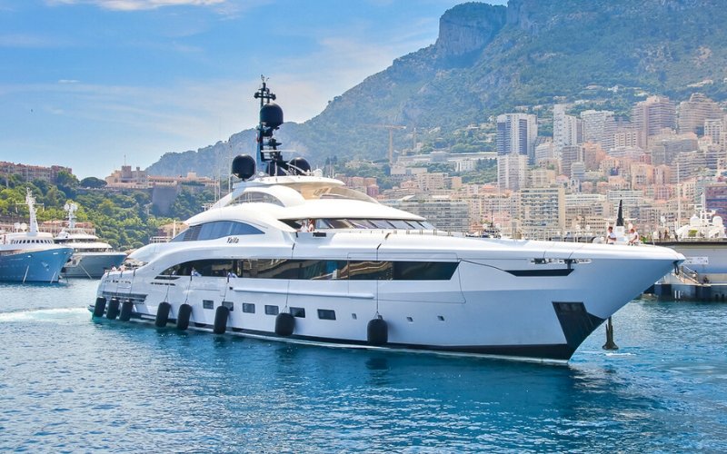 The side of an anchored white super yacht with black fenders along the side, with the sea surrounding it and a coastal area visible in the background