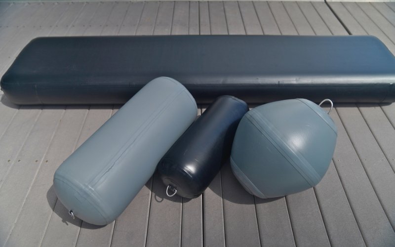Three fenders of varying sizes, two light gray and one black, with a black cushion of the same material behind them, all placed on a wooden deck of a yacht