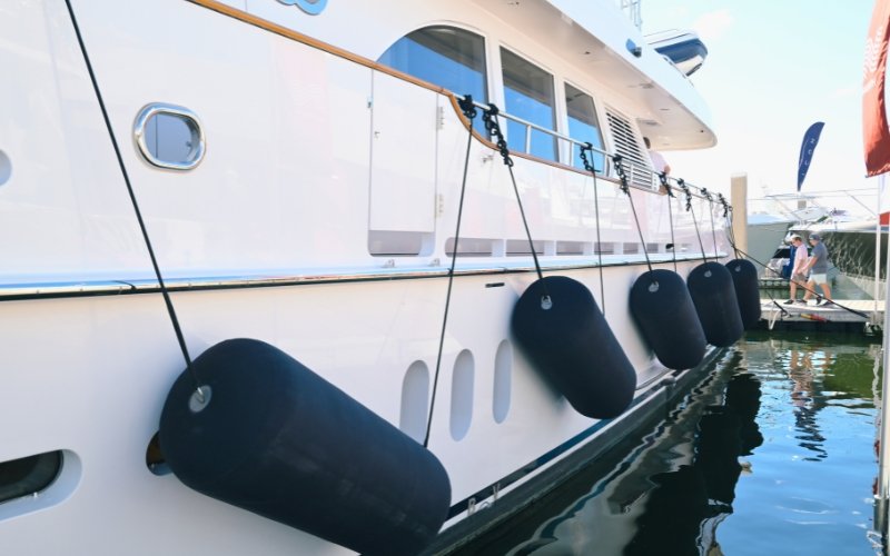 The side of an anchored white super yacht with black fenders along the side, with the sea surrounding it and a coastal area visible in the background