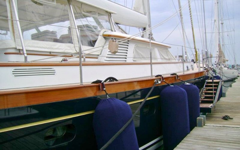 A traditional wooden sailing boat in dark blue and white colors, anchored with three large blue fenders resting against the dock