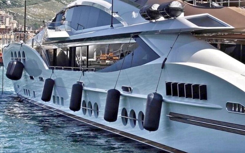 The side of an anchored white super yacht with black fenders along the side, with the sea surrounding it and a coastal area visible in the background