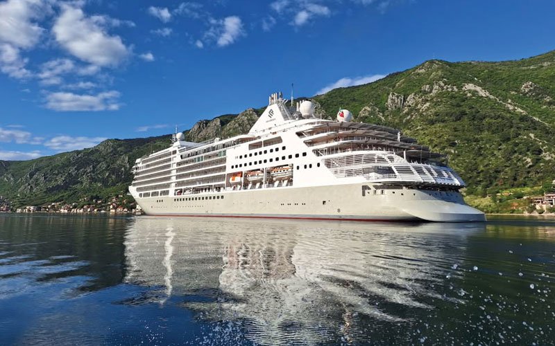 A large cruise ship docked at a port, with reflective sea waters and a lush green landscape in the background