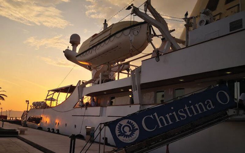 A photograph of the yacht Christina O at sunset, with the golden light reflecting on the water and the yacht silhouetted against the vibrant sky.