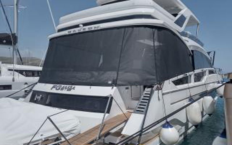A white boat docked in a marina with a grey cover on the front, and a small boat with a white cover placed at the bow. To the right, the dock is visible, and on the side of the boat, the fenders are attached