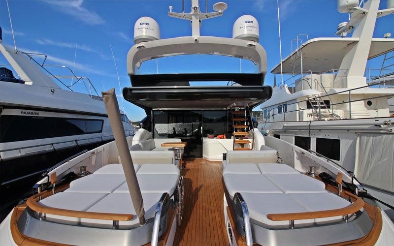 A white yacht with wooden details, a wooden deck, and a wooden ladder, docked in a marina. At the front, there are sunbathing furniture with cushions designed by Argola Textile Yacht Design. In the back, a sofa and lounge area are visible