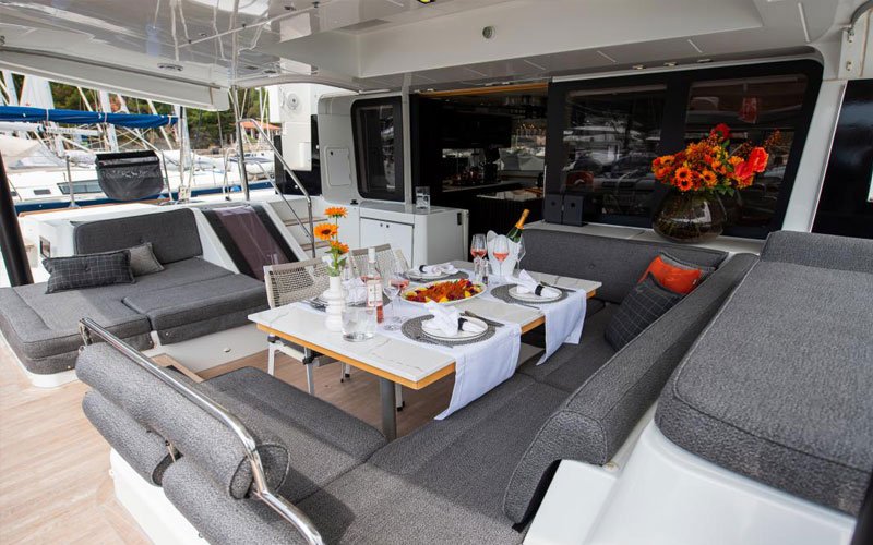 Large grey lounge with a table in the outdoor area of a yacht, anchored in a marina. The table is set with beautiful tableware, food, wine, and champagne