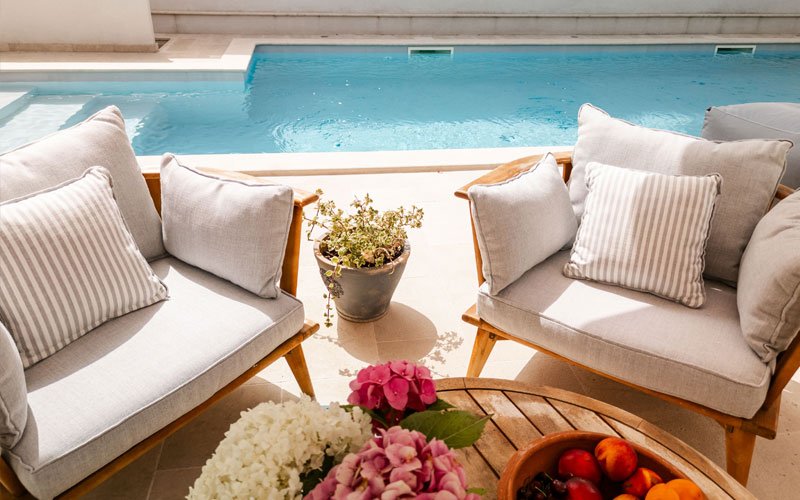 Wooden table with flowers at the front of a yacht deck, two wooden armchairs with cushions designed by Argola Textile Yacht Design, and a pool in the background, creating a sophisticated outdoor space