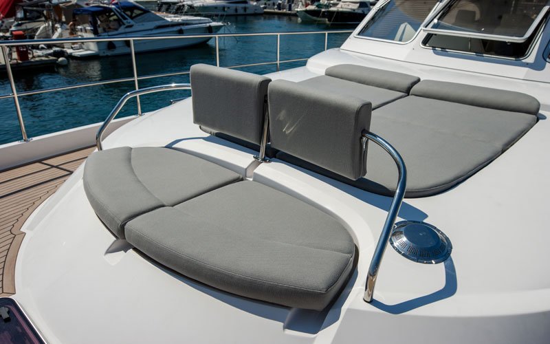 Upper exterior of a yacht anchored in a marina, featuring grey furniture, cushions, and sun loungers for sunbathing, all designed by Argola Textile Yacht Design