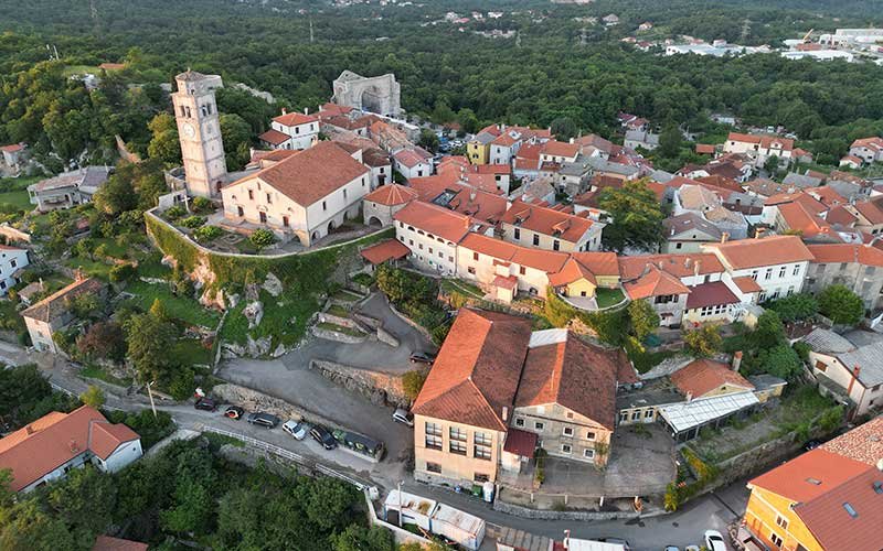 An aerial view of Betina, featuring charming houses, a church, a winding road, and dense greenery in the background