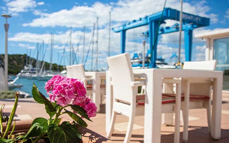 In the foreground, a café and restaurant with a fuchsia flower in front, while the marina and shipyard are subtly visible in the background
