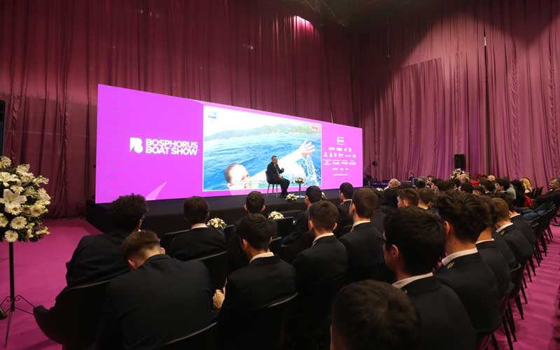 Attendees, including people in formal, are seated in a purple-draped hall at the Bosphorus Boat Show event.