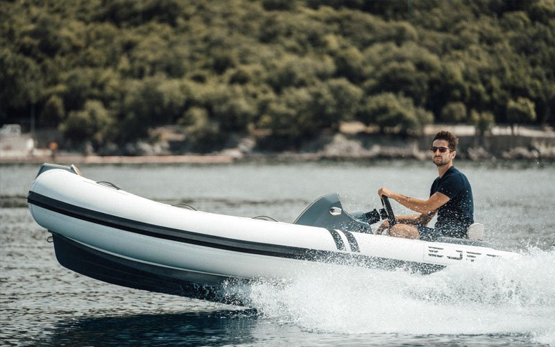 Man driving a jet-powered inflatable boat on the water, with a forested coastline in the background
