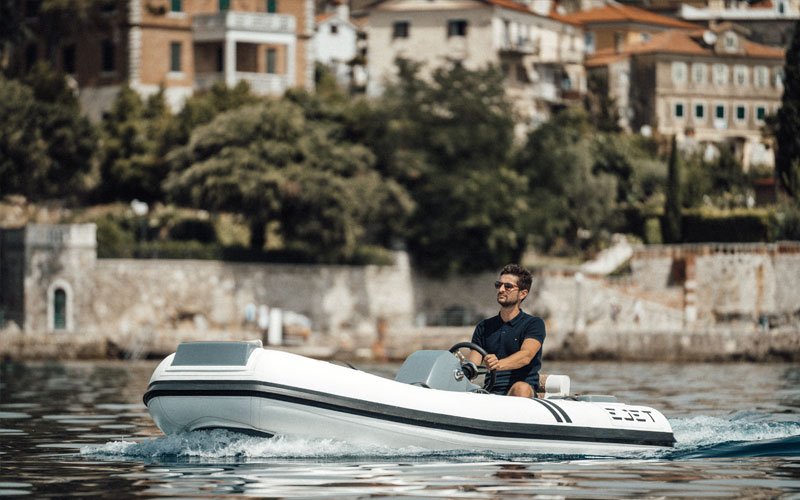 Man driving a jet-powered inflatable boat on the water, with a forested coastline in the background