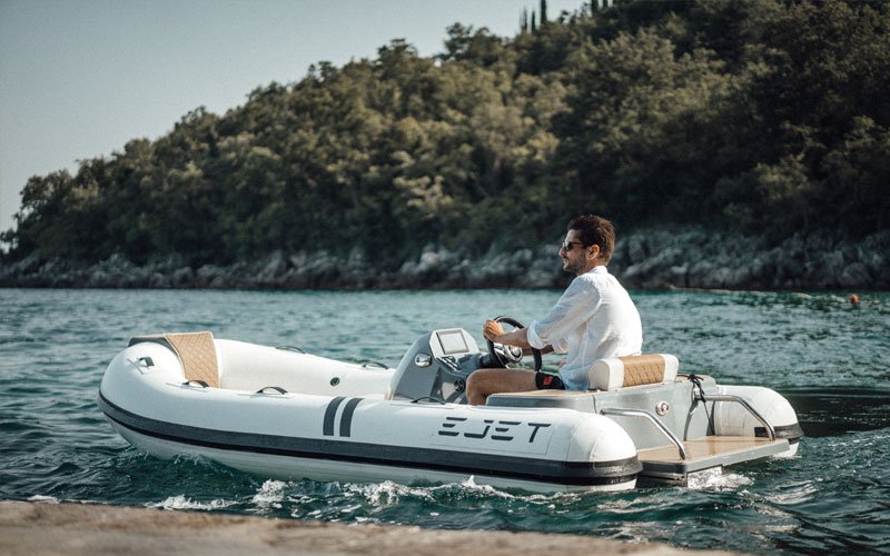 Man driving a jet-powered inflatable boat on the water, with a forested coastline in the background