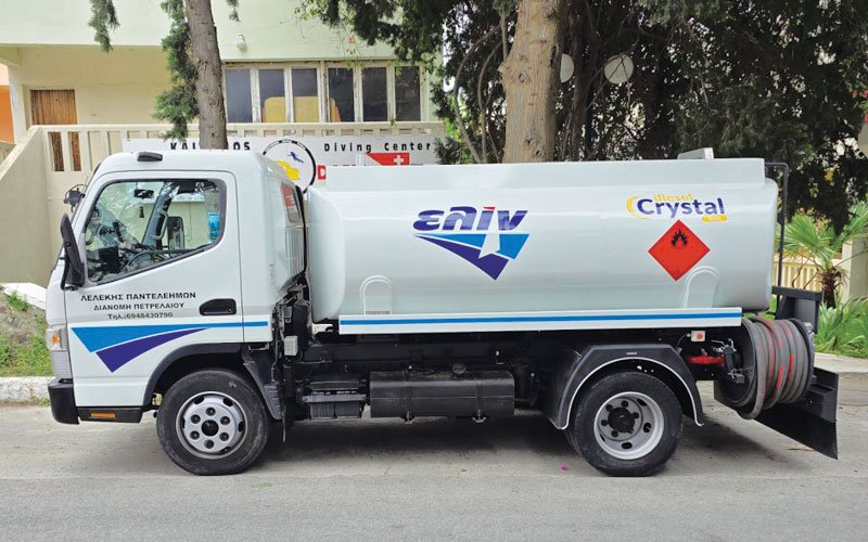 A white fuel delivery truck branded with the ELIN logo and 'Crystal diesel' parked on the street. The vehicle has Greek text on the door indicating petroleum distribution services, along with a visible fuel hose reel at the rear. A diving center building is in the background among trees