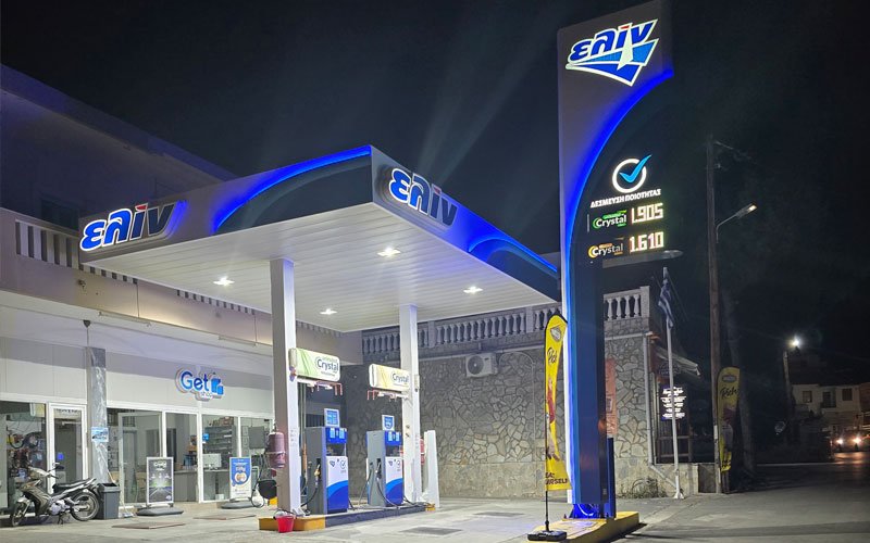 A fuel station branded with 'ΕΛΙΝ' (ELIN), a Greek petroleum company. The station includes a canopy over the pumps, a price sign showing fuel rates, and a white ELIN fuel truck parked nearby.