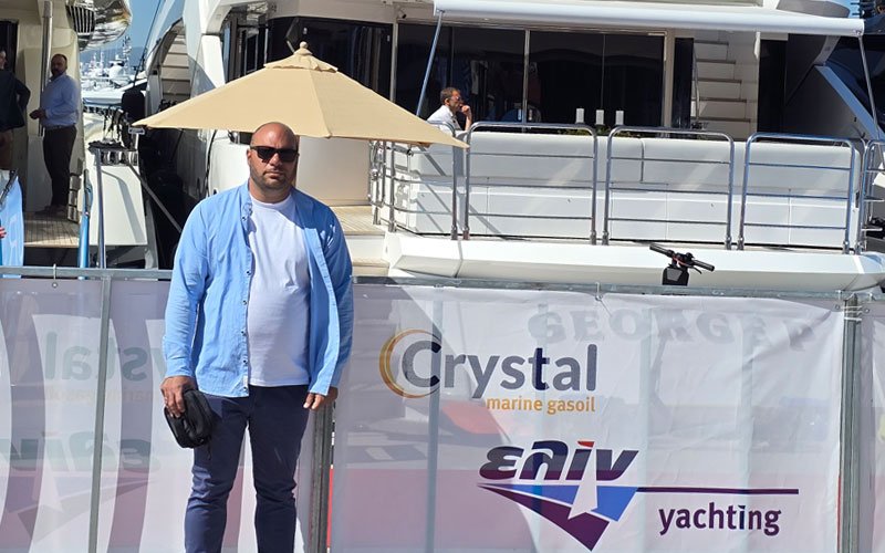 A man wearing sunglasses, a light blue shirt, and dark pants stands in front of a luxury yacht at a marina. Behind him is a banner displaying the logos of “Crystal marine gasoil” and “Eniv yachting.” Several people are visible on the yachts in the background under sunny weather.