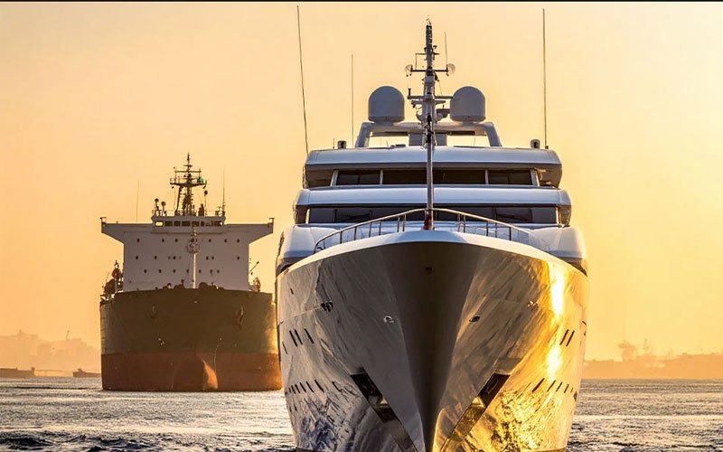 Front view of a luxury superyacht sailing at sunrise with a large cargo vessel in the background, representing modern maritime engineering.