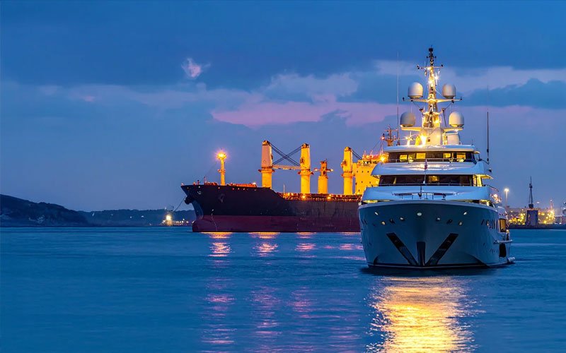Illuminated superyacht navigating a harbor at dusk with industrial cargo ships and port cranes in the background.