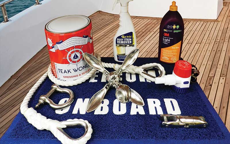 Marine maintenance products displayed on a boat deck, including Teak Wonder Dressing & Sealer, Star brite Rust Stain Remover, 3M Cleaner & Wax, a folding anchor, rope with stainless steel thimbles, shackles, a hose fitting, and a cleat, all placed on a blue welcome mat.