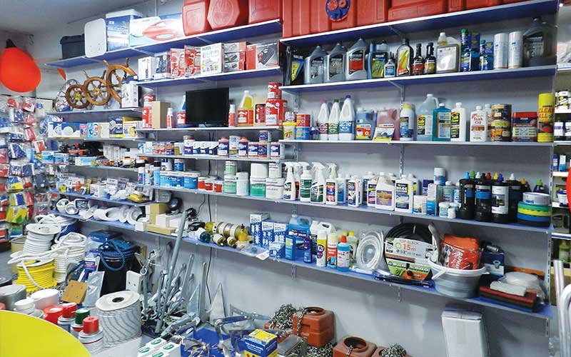 Interior of the LIANOS NIKOLAOS store showcasing a wide range of marine supplies, including ropes, paints, cleaning products, lubricants, flags, boat steering wheels, and various marine accessories neatly organized on shelves.