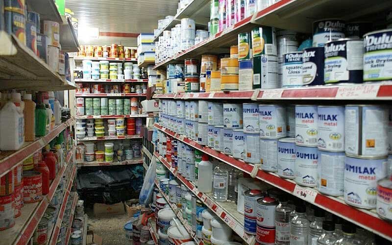 Aisle inside the LIANOS NIKOLAOS store displaying a large variety of paint cans, primers, varnishes, and solvents from brands like Dulux and Gumilak, neatly arranged on shelves along both sides.