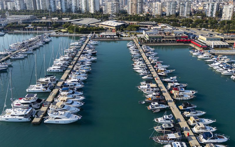 Aerial view of Mersin Marina in Turkey, showing yachts and boats docked in the harbor, surrounded by breakwaters, with the city skyline in the background
