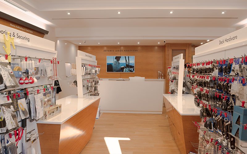 Interior view of Naval Store Yacht Supplies by Artemis Leros Boatyard, featuring light wood furniture and various products on display. In the background, the reception area with a monitor visible behind it