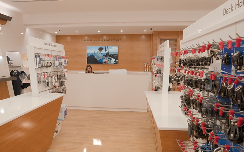 Interior view of Naval Store Yacht Supplies by Artemis Leros Boatyard, featuring light wood furniture and various products on display. In the background, the reception area with a monitor visible behind it