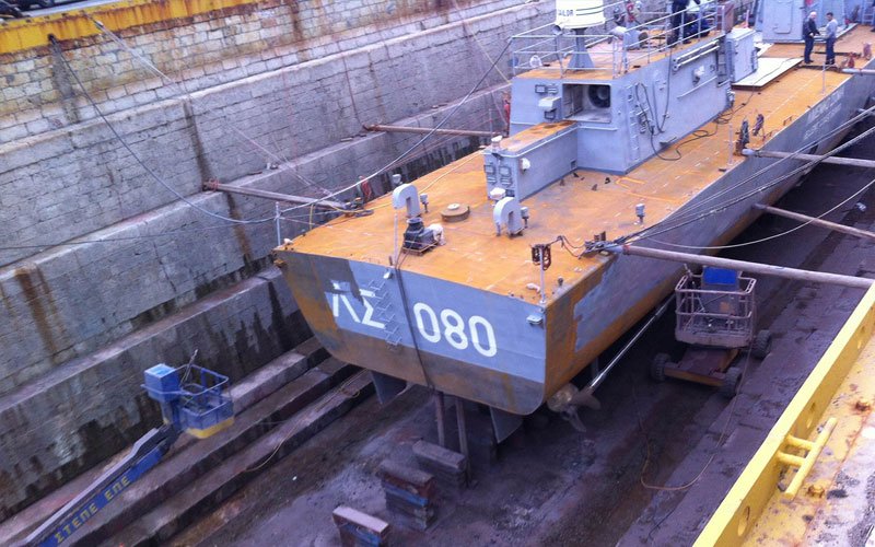 A military vessel with the number 080 painted on its stern is in a dry dock undergoing maintenance, with workers and equipment visible around it