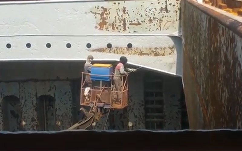 Two workers in protective gear on a lift platform cleaning or sandblasting the rusted hull of a large ship in a dry dock.