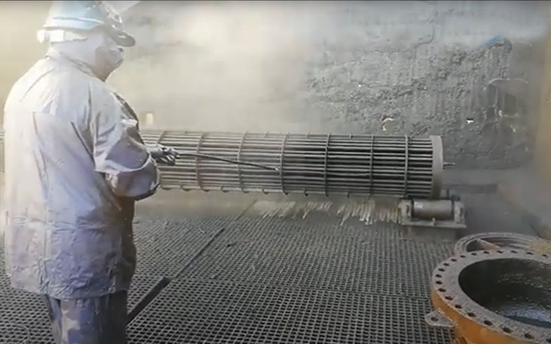 Worker in protective gear using a high-pressure hose to clean or sandblast a large cylindrical metal heat exchanger inside an industrial facility
