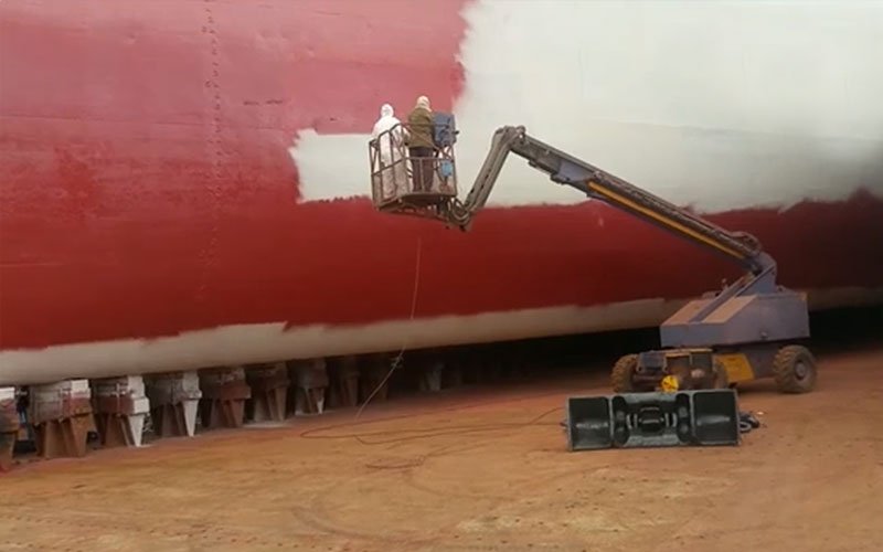 Two workers in protective gear on a lift platform painting the rusted hull of a large ship in a dry dock.