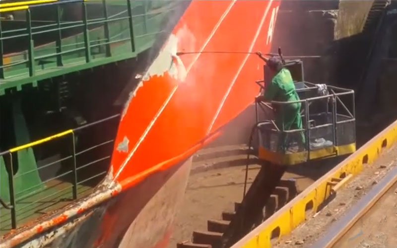 A worker wearing protective gear uses a high-pressure washer to clean or strip paint from the hull of a large ship in a dry dock.