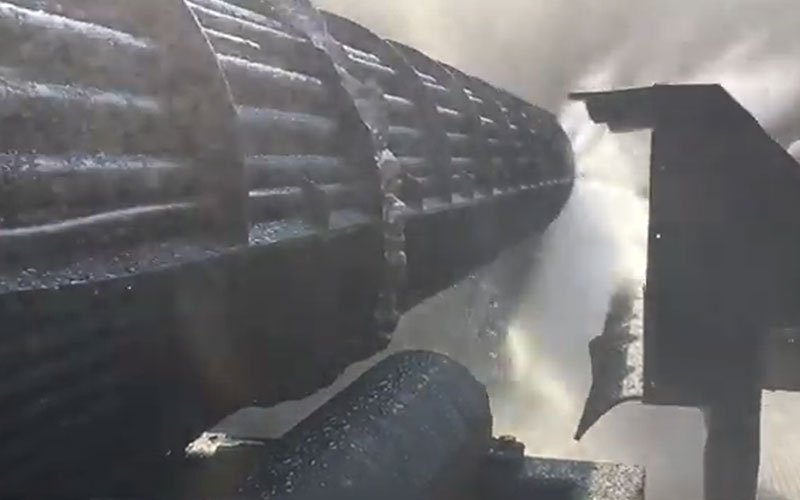 Close-up view of a large industrial roller or drum being cleaned with high-pressure water jets, with water spraying and mist surrounding the surface.