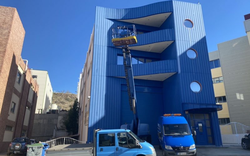 A blue telescopic boom lift extends from a blue utility truck, lifting a work platform up the side of a modern blue building with circular windows. The platform is positioned near the top floor of the building, possibly for maintenance or construction work. Several other vehicles are parked nearby.