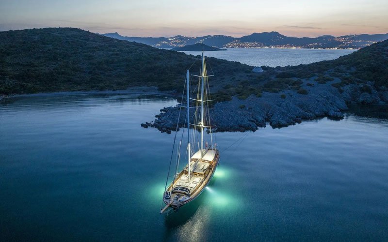 Large sailboat illuminated by stunning lights, reflecting on the calm sea at dusk, with a warm, colorful sky in the background.