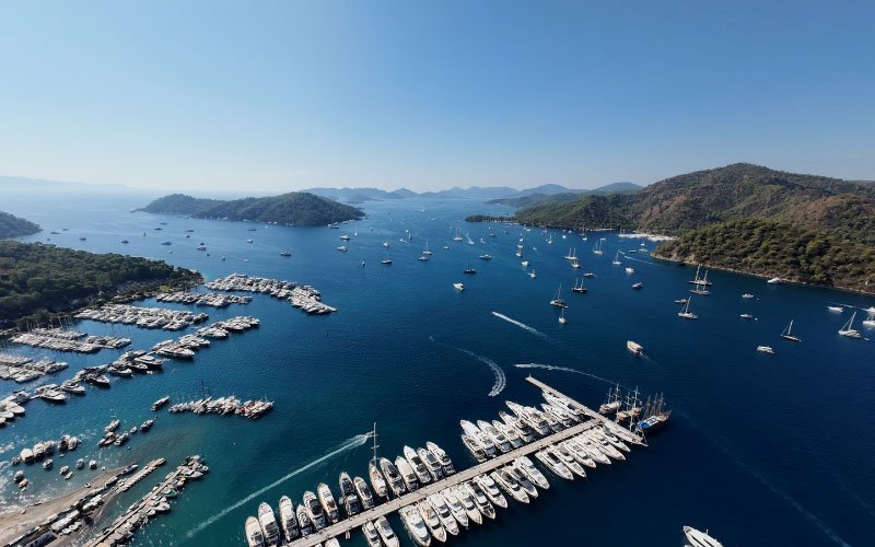 Docked boats in a marina, with additional vessels anchored both inside and outside the bay, floating on clear blue waters