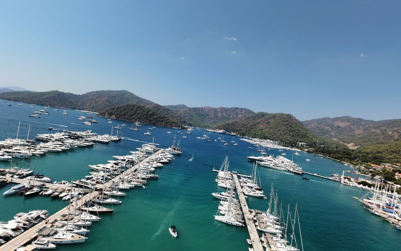 Docked boats in a marina, with additional vessels anchored both inside and outside the bay, floating on clear blue waters