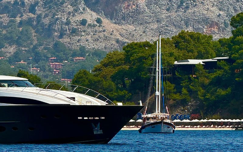 The bow of a mega yacht with a sailing yacht positioned perpendicular to it, set against a backdrop of a beach with umbrellas and sun loungers, surrounded by lush green scenery