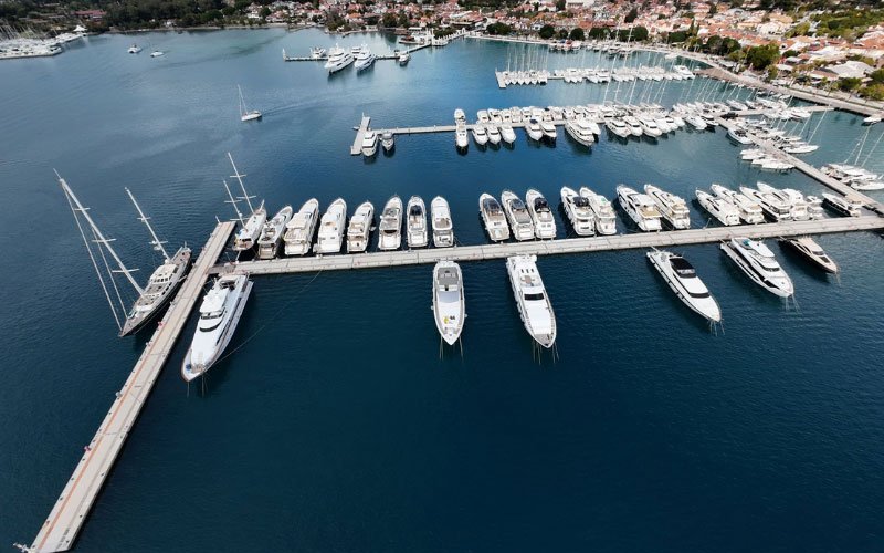 Docked boats in a marina, with additional vessels anchored both inside and outside the bay, floating on clear blue waters