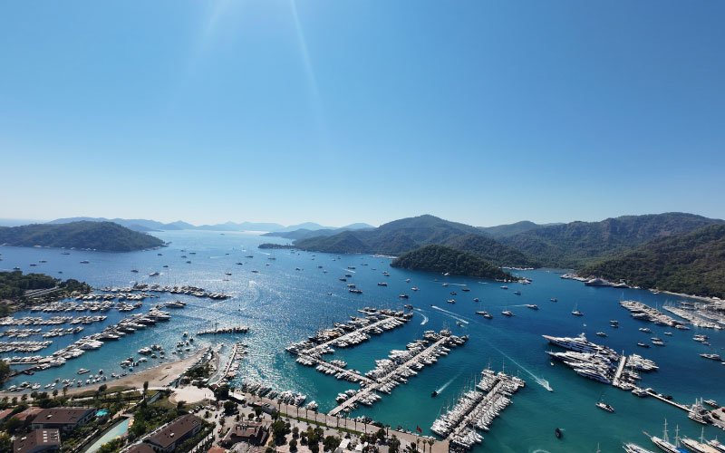 Docked boats in a marina, with additional vessels anchored both inside and outside the bay, floating on clear blue waters
