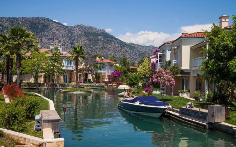 A picturesque scene featuring a canal surrounded by trees and palm trees, with charming traditional houses in the background and a speedboat moored along the side