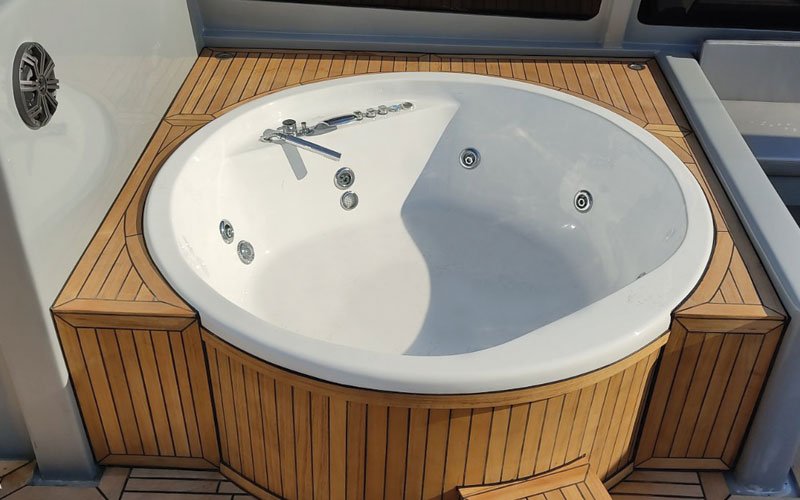 A round white jacuzzi with built-in jets and a faucet, surrounded by a wooden deck with a marine-style finish. The jacuzzi is installed on a boat or yacht, with a speaker mounted on the adjacent white wall.