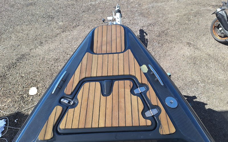 The bow of a boat featuring a wooden deck with a synthetic teak finish, black trim, and metal hardware. A hatch with a black frame is integrated into the deck, along with cleats and other fittings.