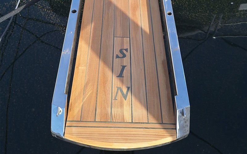 A wooden boat swim platform with a synthetic teak finish, bordered by polished stainless steel rails. The word "SIN" is engraved or embedded in the center. The platform extends over the water, reflecting sunlight