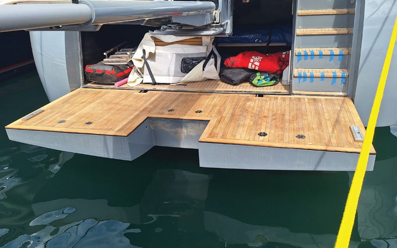 A close-up view of the stern of a yacht named "VAKOMAR" registered in London. The yacht features a teak wood deck with a hatch in the center, metal fittings, and a stainless-steel handrail.