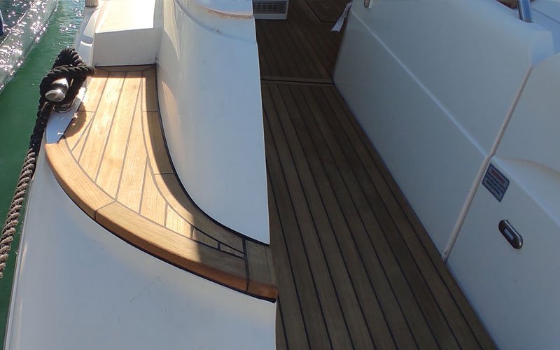 A close-up view of a yacht's teak deck, showing a curved wooden step with a polished finish. A thick black mooring rope is secured on the left side near a metal cleat.