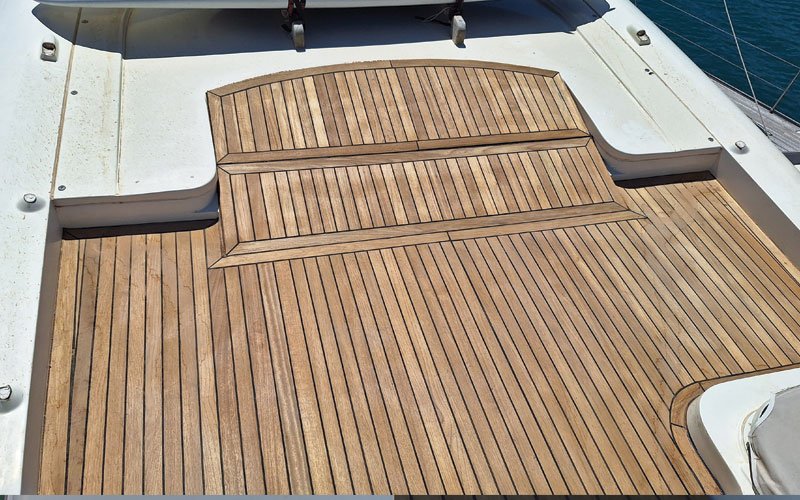 A close-up of a yacht's teak deck, featuring a raised, curved step with a symmetrical plank pattern. The deck has a warm, natural wood tone with visible grain and black caulking lines.